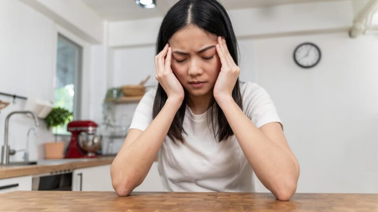 a person with long black hair and a white tee holding their head with their hands with elbows resting on a wood counter in a kitchen.