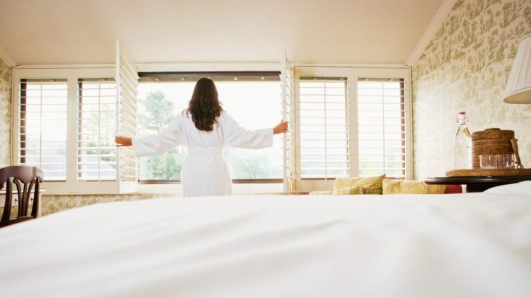 A brunette woman in a white fluffy bathrobe opens white shutters to let sunlight into her rustic European hotel room.