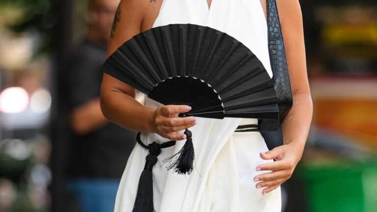 Woman in a sleeveless white dress holds a black handheld fan with tassel outside Viktor & Rolf during Paris Fashion Week