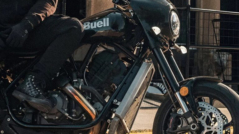 Close-up of a black Buell motorcycle focusing on the engine, front wheel, and part of the fuel tank with the Buell logo. The rider is wearing black pants, black gloves, and black checkered boots. The motorcycle has a round headlight and visible front disc brakes.