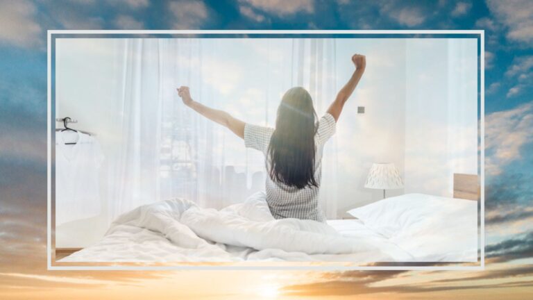 A woman sat on edge of white bed stretching her arms overhead looking out to morning sun