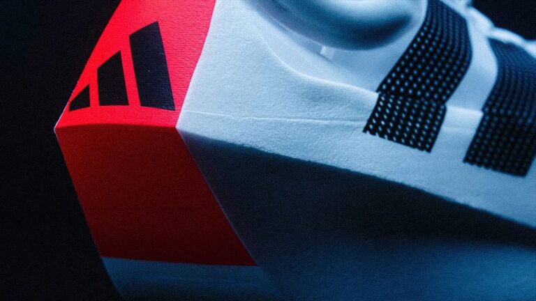 Adidas Can Officially Claim the Boldest Super Shoe of the Year and It’s Not Close Close-up of the heel area of a white Adidas sneaker with black stripes and a red section featuring the Adidas logo. The shoe material appears textured, and the image has a cool blue lighting effect.