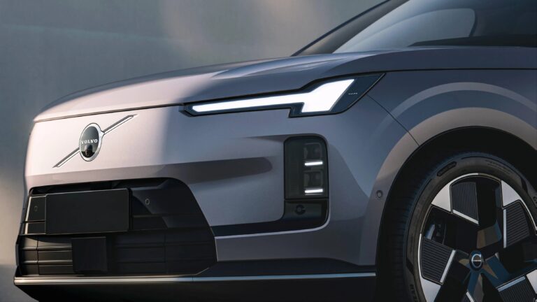 Front side view of a modern Volvo car in gray, featuring a sleek, illuminated headlight design, a smooth front grille with the Volvo logo, and a stylish alloy wheel with a unique pattern.