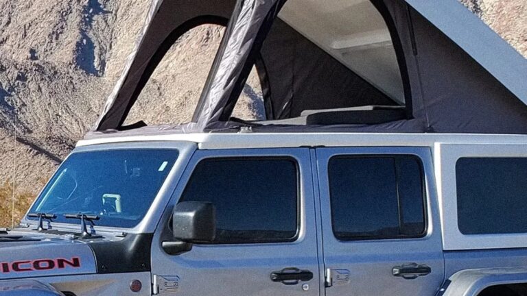Silver Jeep with a black pop-up roof tent extended, parked in a rocky desert area. The Jeep has tinted windows and the word