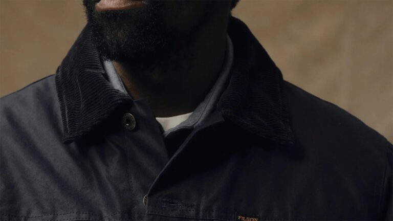 Close-up of a person wearing a dark jacket with a corduroy collar, layered over a gray hoodie and a white shirt underneath. The jacket has a small "FILSON" label on the chest. The person