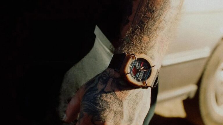 A close-up of a wrist wearing a watch with a wooden bezel and a dark face featuring multiple dials. The wrist has a detailed tattoo, and the arm is covered with light-colored hair. The watch strap appears to be dark, possibly black or brown. The background is blurred and dimly lit.