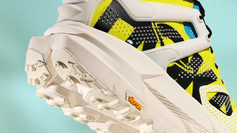 Chunky sneaker with a thick, textured off-white sole featuring deep treads and a small orange Vibram logo. The upper part is bright yellow with black and blue accents, and has a white supportive frame around the heel and sides. The shoe has a modern, sporty design with a mix of mesh and synthetic materials.