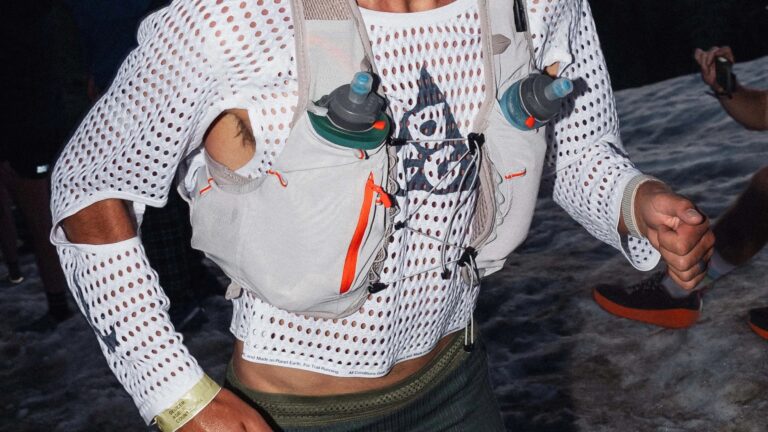 Close-up of a person wearing a white, long-sleeve, perforated mesh shirt with a black logo on the chest. They have a light gray hydration vest with two water bottles secured in front pockets, featuring orange zipper accents and adjustable cords. The person is running, with a gold wristband on their left wrist and dark green shorts visible at the waist. The background shows a dark, outdoor setting with water and other people partially visible.