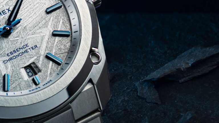 Close-up of a silver formex wristwatch with a textured dial featuring blue hour markers and hands. The dial includes the text