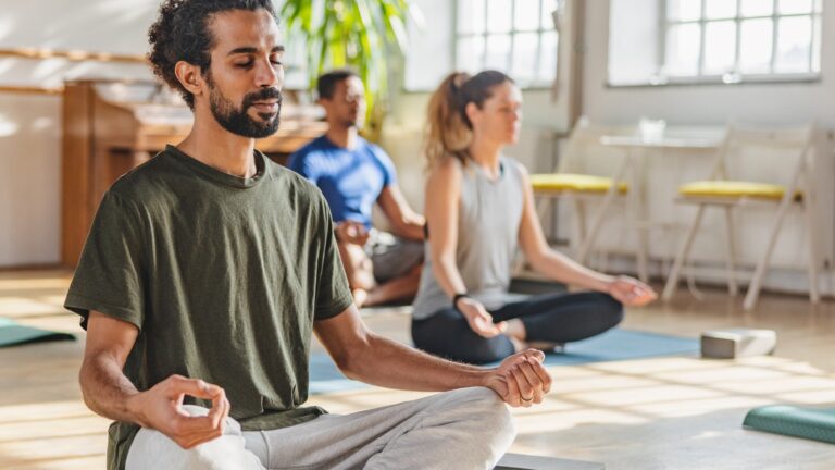 A man with a ponytail meditates in a yoga class, surrounded by other yoga class participants in a bright studio with large windows and plants.