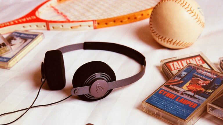 A pair of black and gray over-ear headphones lies on a white surface. Nearby are several cassette tapes, including one with a cover featuring Cyndi Lauper
