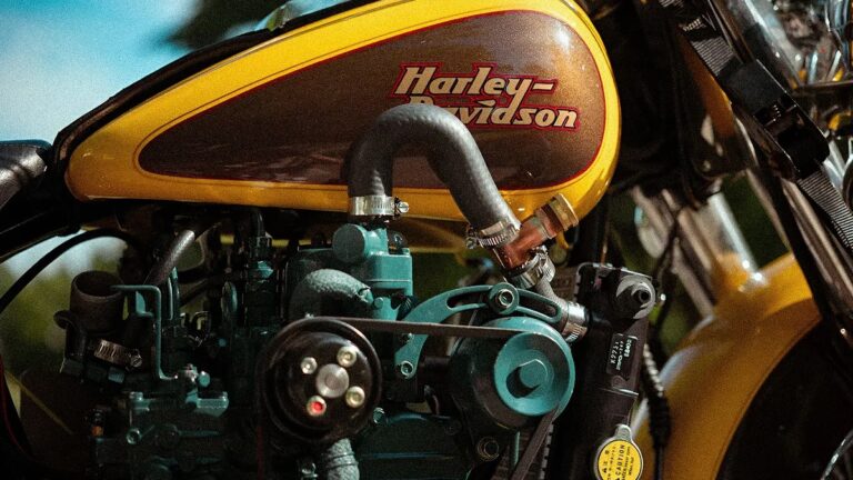Close-up of a Harley-Davidson motorcycle fuel tank and engine, featuring a yellow and brown fuel tank with the Harley-Davidson logo, black hoses, and various engine components in teal and black.