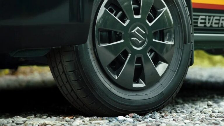 Close-up of a black car tire with a Suzuki logo on the hubcap, resting on a gravel surface. The car