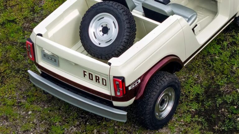 Rear view of a cream and maroon Ford Bronco with a spare tire mounted in the truck bed, parked on grass. The tailgate displays the