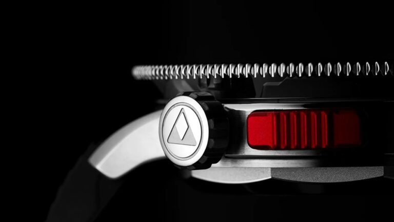 Close-up of an albishorn watch crown with a triangular logo, a textured bezel, and a red rectangular button on the side, set against a black background.