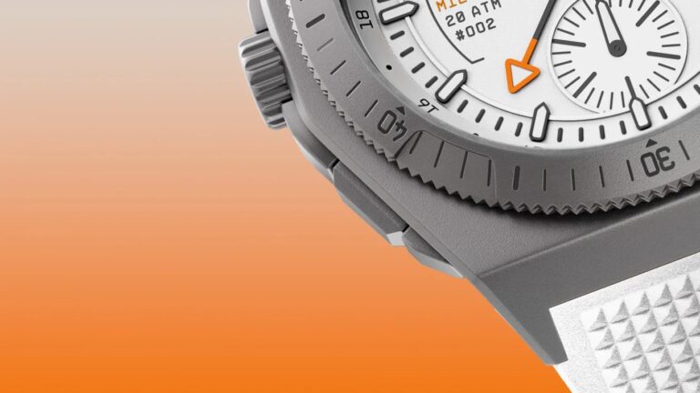 Close-up of a gray micromilspec wristwatch with a textured bezel marked with minute indicators, a white dial featuring black hour markers, an orange arrow-shaped hand, and a subdial. The watch case has a matte finish and a textured white strap. The background is a gradient from light beige to orange.