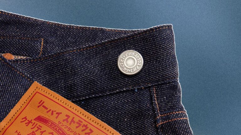 Close-up of the waistband of dark blue denim jeans with orange stitching, featuring a metal button with Japanese characters and a tan leather patch with red Japanese text.