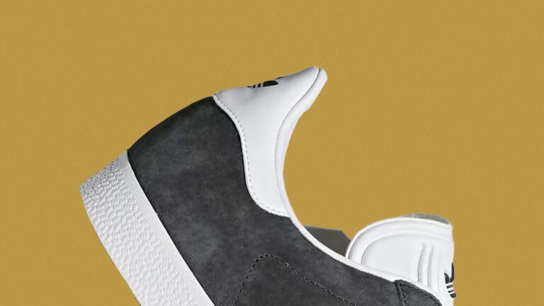 Close-up of a sneaker featuring a dark gray suede upper with white leather accents around the heel and ankle collar, and a textured white rubber sole. The background is a solid mustard yellow color.