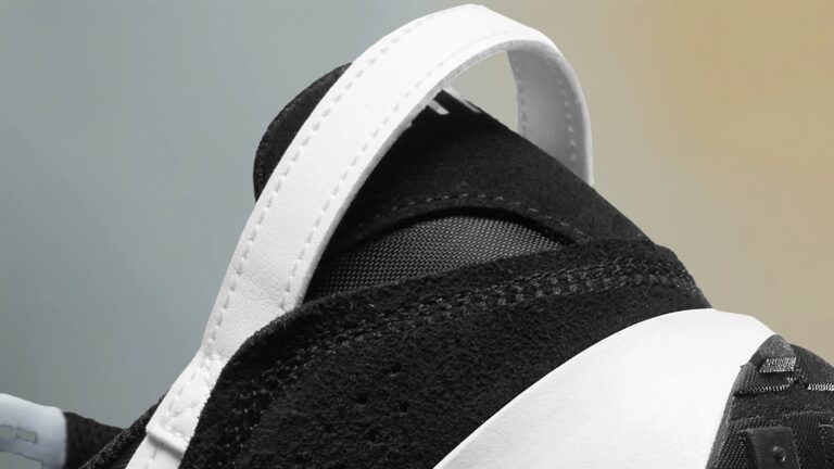 Close-up of the back heel area of a black and white sneaker, featuring a white pull tab with visible stitching and a textured black upper with mesh detail. The midsole is white, and part of the black outsole is visible.