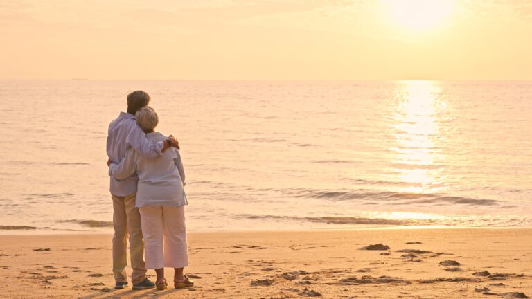 An older couple stand arm in arm on a beach and watch the sunset.