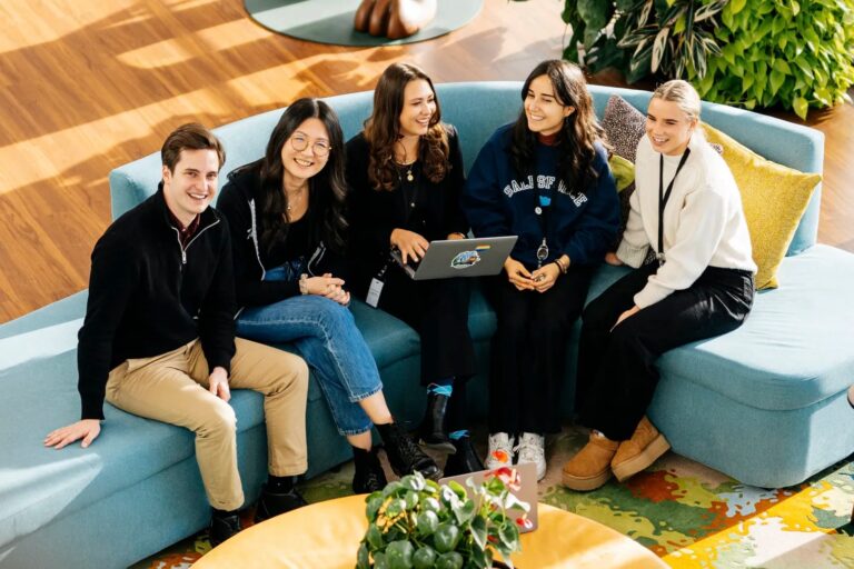 A group of five young professionals sits together on a blue couch, smiling and collaborating around a laptop in a bright, modern office space.