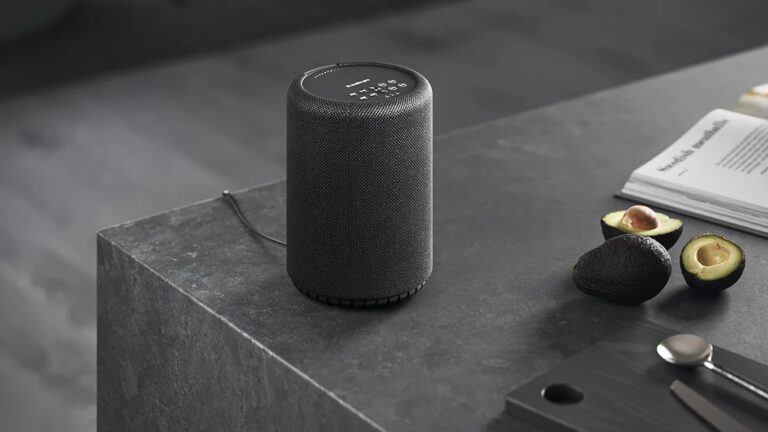A cylindrical black smart speaker with a fabric exterior sits on a dark gray stone countertop. Next to it are two halved avocados, one whole avocado, an open book, and a black cutting board with a spoon and knife.
