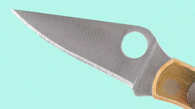 Close-up of a knife blade with a sharp edge and a circular hole near the handle, which is made of wood with a visible grain pattern. The background is a solid light turquoise color.