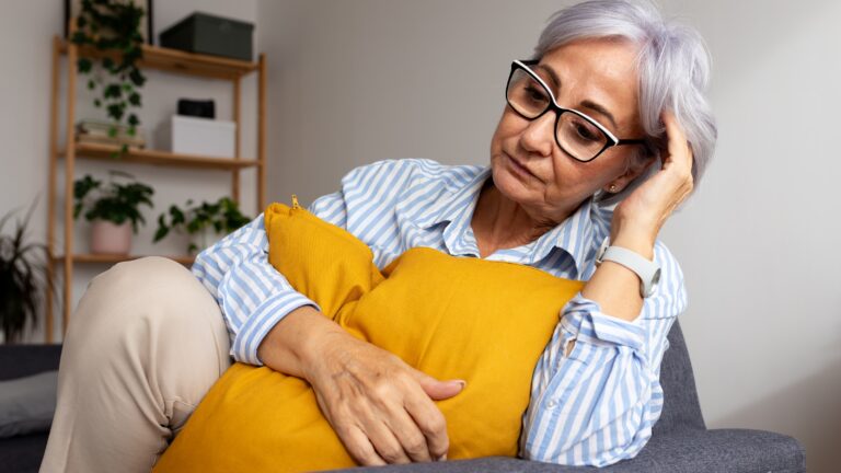 How to Navigate Your Finances After Losing Your Spouse An older woman looks serious as she sits on her sofa, holding a pillow to her chest.