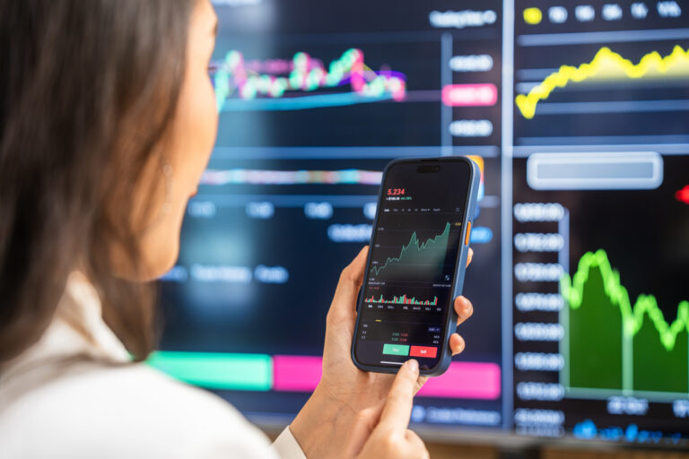 I’m 55 With 10 Years Until Retirement and I’ve Made $2 Million on Nvidia Stock. What Do I Do With It Now? A businesswoman examines financial charts and graphs on her smartphone, utilizing modern technology for investment analysis amidst digital screens displaying stock data.