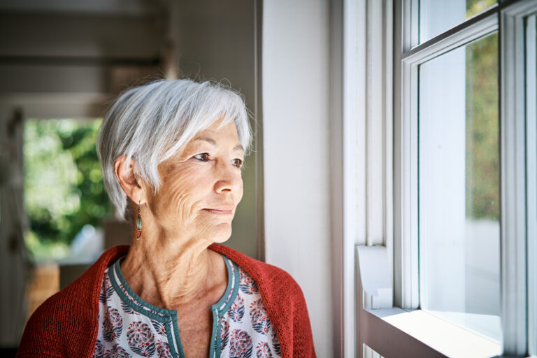 I’m a 76-Year-Old Widow and My Son Is Pushing Me Into Assisted Living. How Do I Convince Him I’m Fine Living on My Own? Senior woman standing alone in her living room at home and looking out through a window