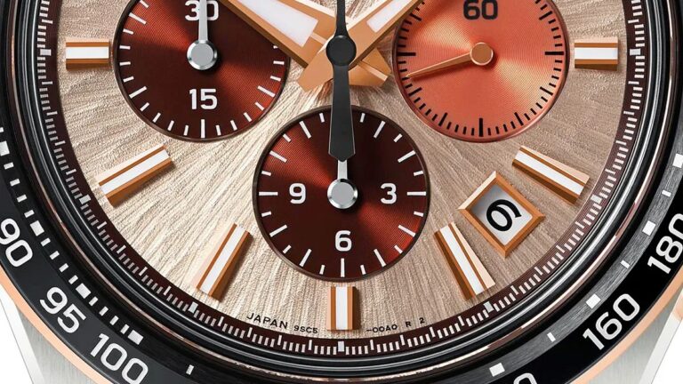 Is This Grand Seiko’s Most Impressive Chronograph Yet? Close-up of a watch face featuring a textured rose gold background with three subdials in a darker copper tone. The subdials display 30, 60, and 6 markings. The main dial has rectangular hour markers with white and rose gold accents. A date window shows the number 6, framed in rose gold. The outer bezel is black with white numeric markings ranging from 90 to 180. The watch hands are rose gold with white inlays, and the second hand is black. Small text near the bottom reads