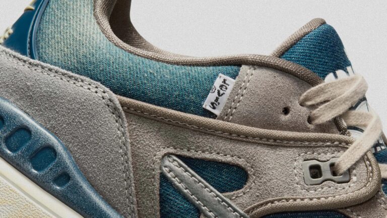 Close-up of a sneaker showing a combination of blue woven fabric and light gray suede materials. The shoe features beige laces, detailed stitching, and a small white tag with black text. The midsole is off-white with a blue plastic support element.