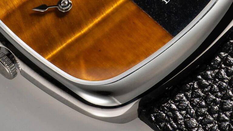 One of the World’s Hottest Affordable Watches Is Now Literally Twice As Nice Close-up of a Dennison wristwatch corner showing a polished silver metal case, a brown stone dial, and a black textured leather strap. The watch crown is partially visible on the left side.