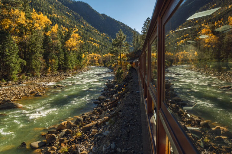 Seven Gorgeous Train Trips to Enjoy Fall Foliage A river and a mountain scene reflect in the window of a passenger railroad car going through an autumn scene. Photo taken October 15 2022 near Durango Colorado, USA.