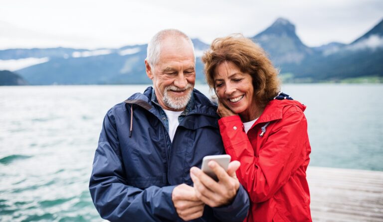 The 10 Best Travel Hacks Every Active Retiree Should Know A mature couple on vacation in a beautiful, mountain location on the water look at a cell phone.