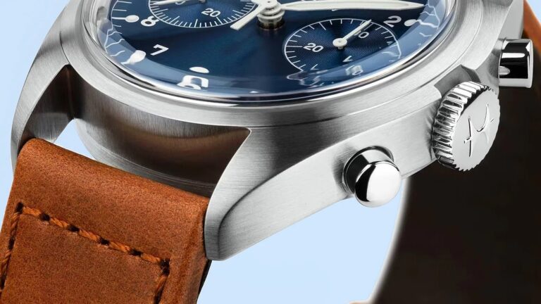 Close-up of a wristwatch featuring a brushed stainless steel case, a blue dial with white numerals and subdials, and a brown leather strap with visible stitching. The watch has two polished pushers and a crown with an engraved logo. The crystal is slightly domed, reflecting light.