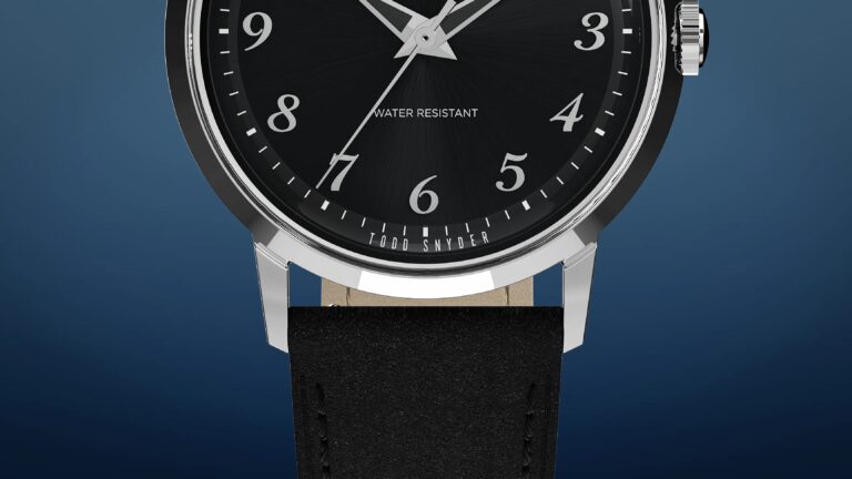 Close-up of a wristwatch with a black dial featuring silver Arabic numerals and hands. The watch face includes the text