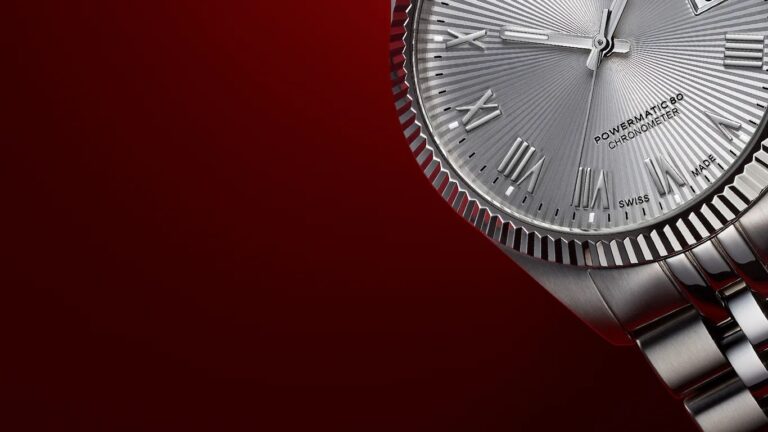 Close-up of a silver Tissot wristwatch with a fluted bezel and a metal link bracelet. The watch face features Roman numeral hour markers, a textured radial pattern, and the text