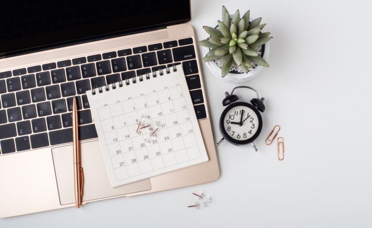 An aerial view of a computer calendar, pen, plant, and time clock to represent time blocking.