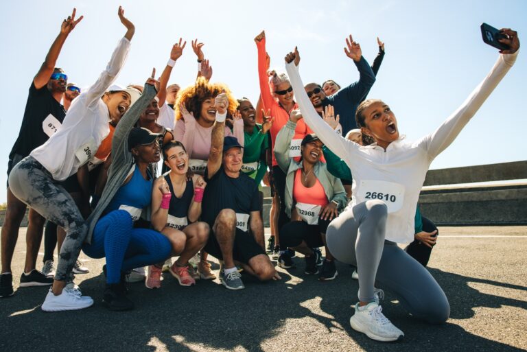 A diverse group of runners celebrates by taking a joyful selfie after completing a race. They're dressed in athletic wear with race numbers, capturing a moment of triumph and unity on a sunny day.